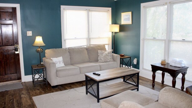 Una sala de estar con un sofá color beige, una mesa de centro y una lámpara. Hay una puerta de madera y una ventana con persianas.