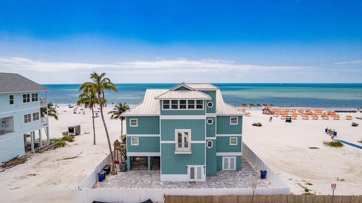 Ein Objekt direkt am Strand mit einem hellblauen Gebäude, einem Sandstrand und blauem Himmel.