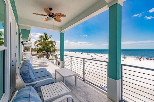 A balcony with lounge chairs and a table overlooking a beach with people and palm trees.