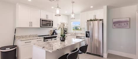 Cocina moderna con armarios blancos, una isla central con taburetes de bar, electrodomésticos de acero inoxidable e iluminación colgante.