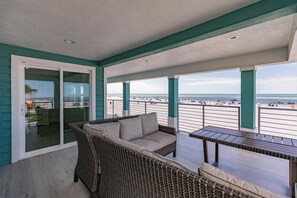 A balcony with a wicker sofa and a table, overlooking a beach with people and umbrellas.