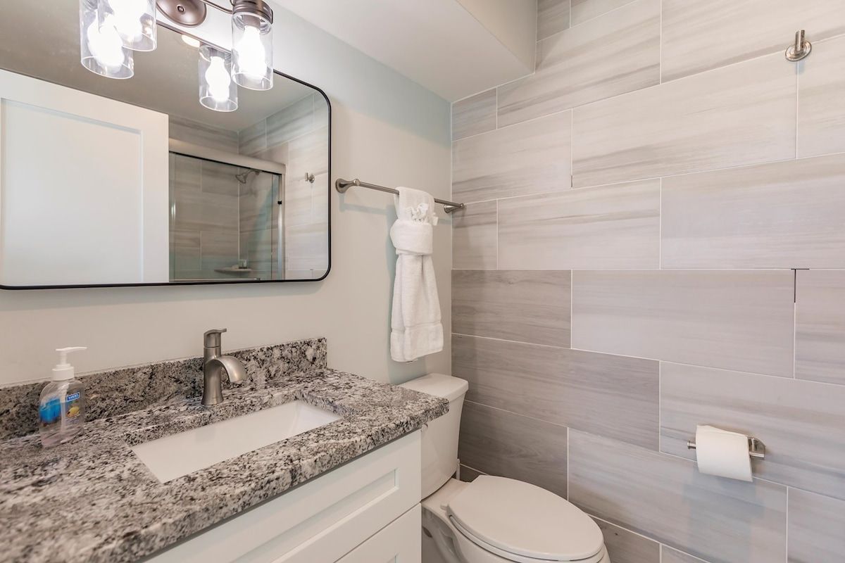 A bathroom with a granite countertop, a white sink, a mirror, a towel rack, and a toilet.