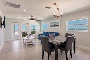 A spacious living area with a dining table, chairs, a ceiling fan, and a view of the ocean through a large window.
