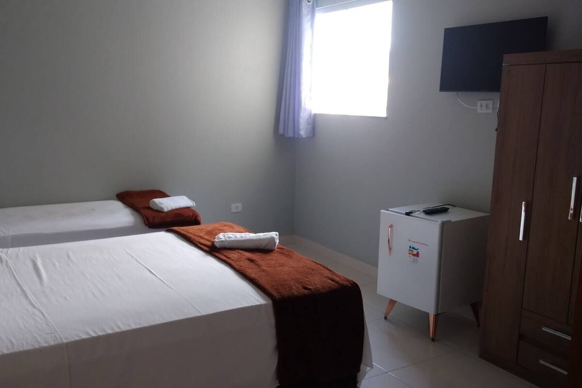 A room with two single beds, a small table with a brown cloth, a white cabinet, and a wardrobe.