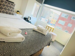 Habitación de hotel con cama, toallas, una ventana con vistas a edificios, una mesita con hervidor y una planta en maceta.