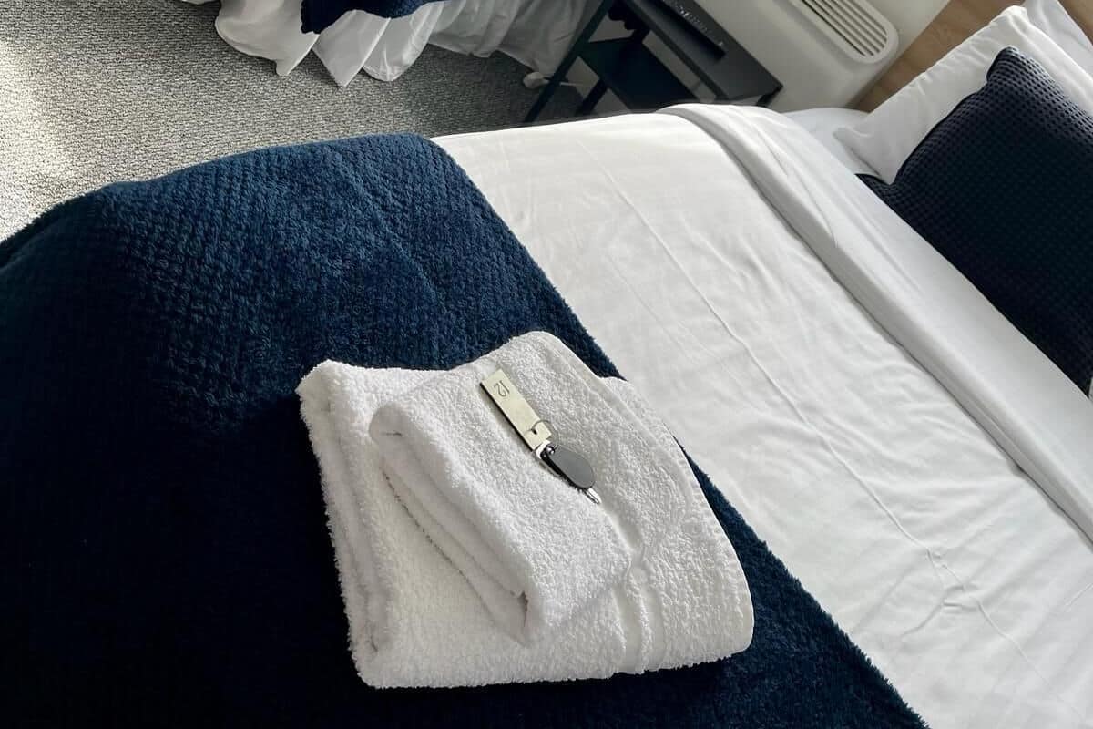 A hotel room with two beds, each with a white mattress and a dark blue blanket. There is a white towel folded on the bed in the foreground.