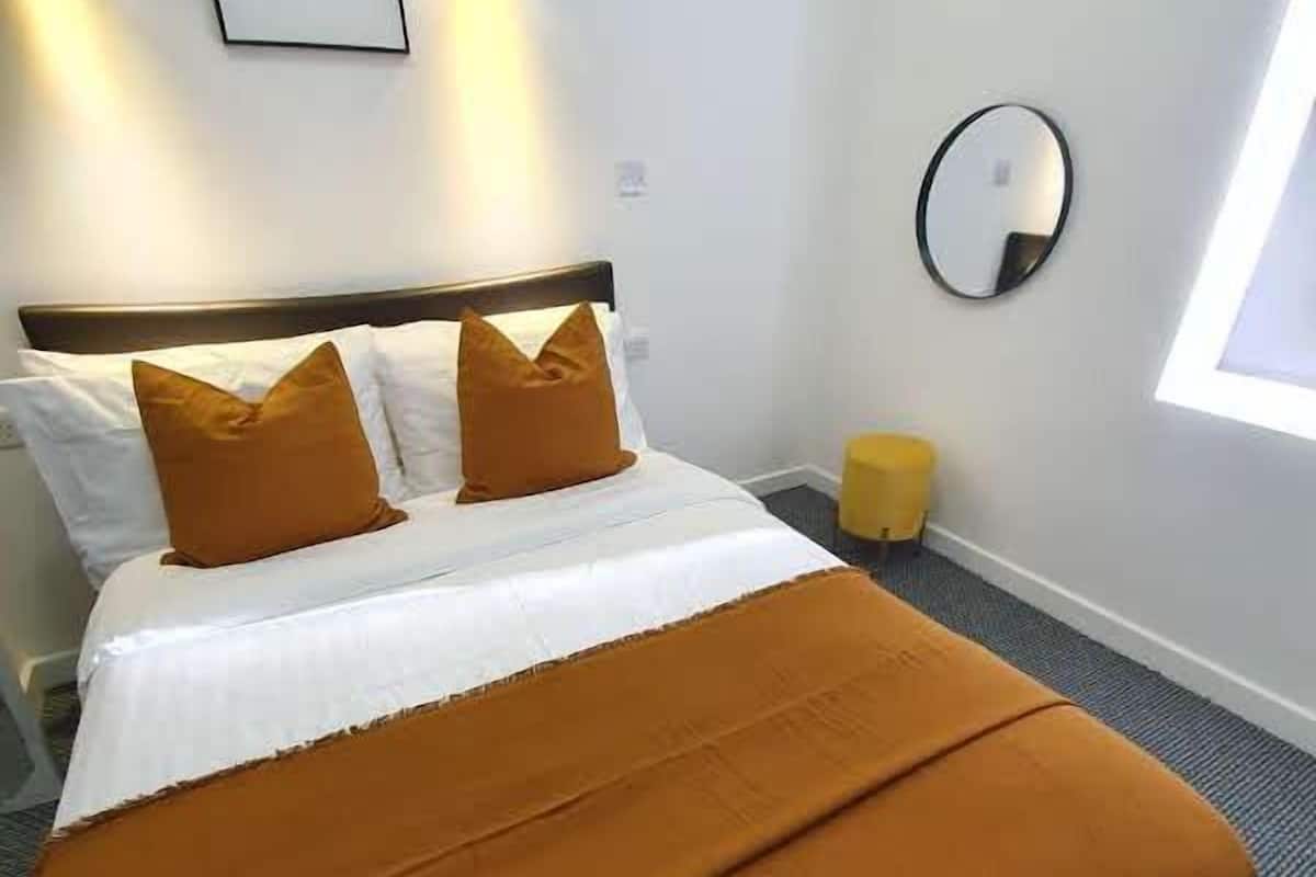 A neatly made bed with a brown bedspread and two matching pillows, a bedside lamp, a round mirror, and a window with daylight.