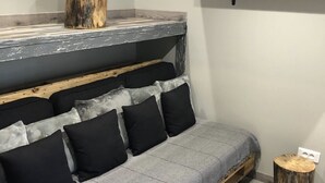 Un canapé rustique agrémenté d’une couverture grise et de coussins noirs, une table d’appoint en bois et une étagère avec des plantes en pot.