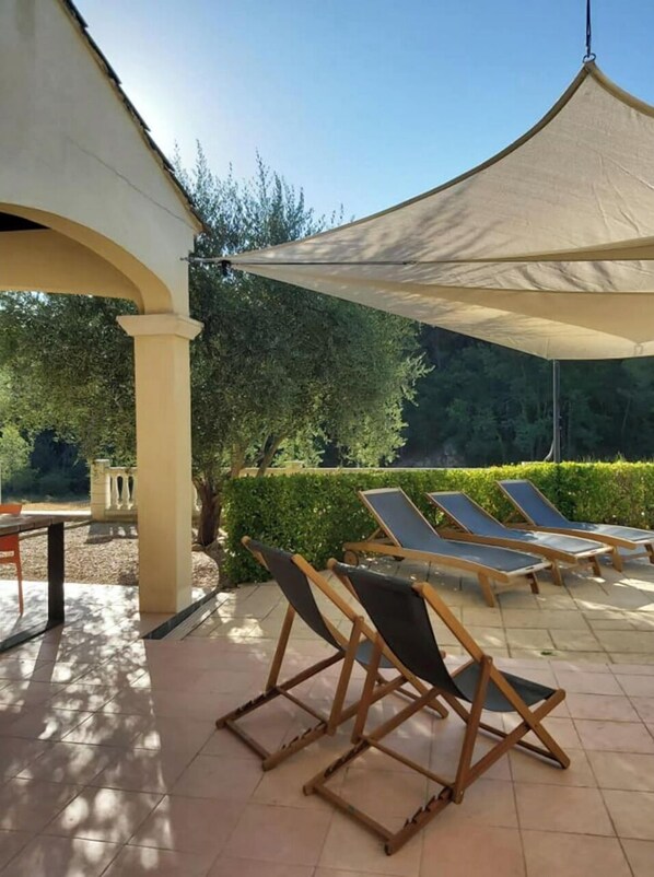 An outdoor seating area with sun loungers under a canopy, surrounded by greenery and a tiled patio.
