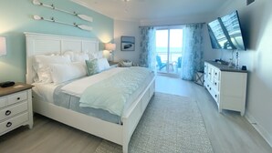 Ein Schlafzimmer mit Bett, Nachttisch, Kommode, Fernseher und Meerblick durch eine Schiebetür.