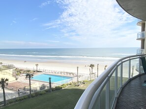 A balcony with a view of a beach, a pool, and a clear sky.