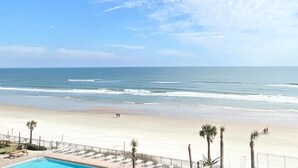 Blick auf einen Strand mit Poolbereich, Palmen und blauem Himmel.