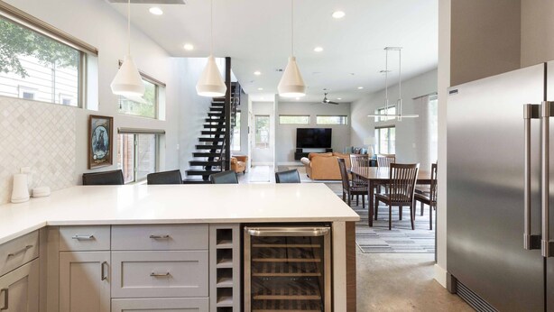 Een moderne keuken met een centraal eiland, hangende verlichting, een ingebouwde wijnkoeler en een eethoek met een trap naar een hoger gelegen verdieping.