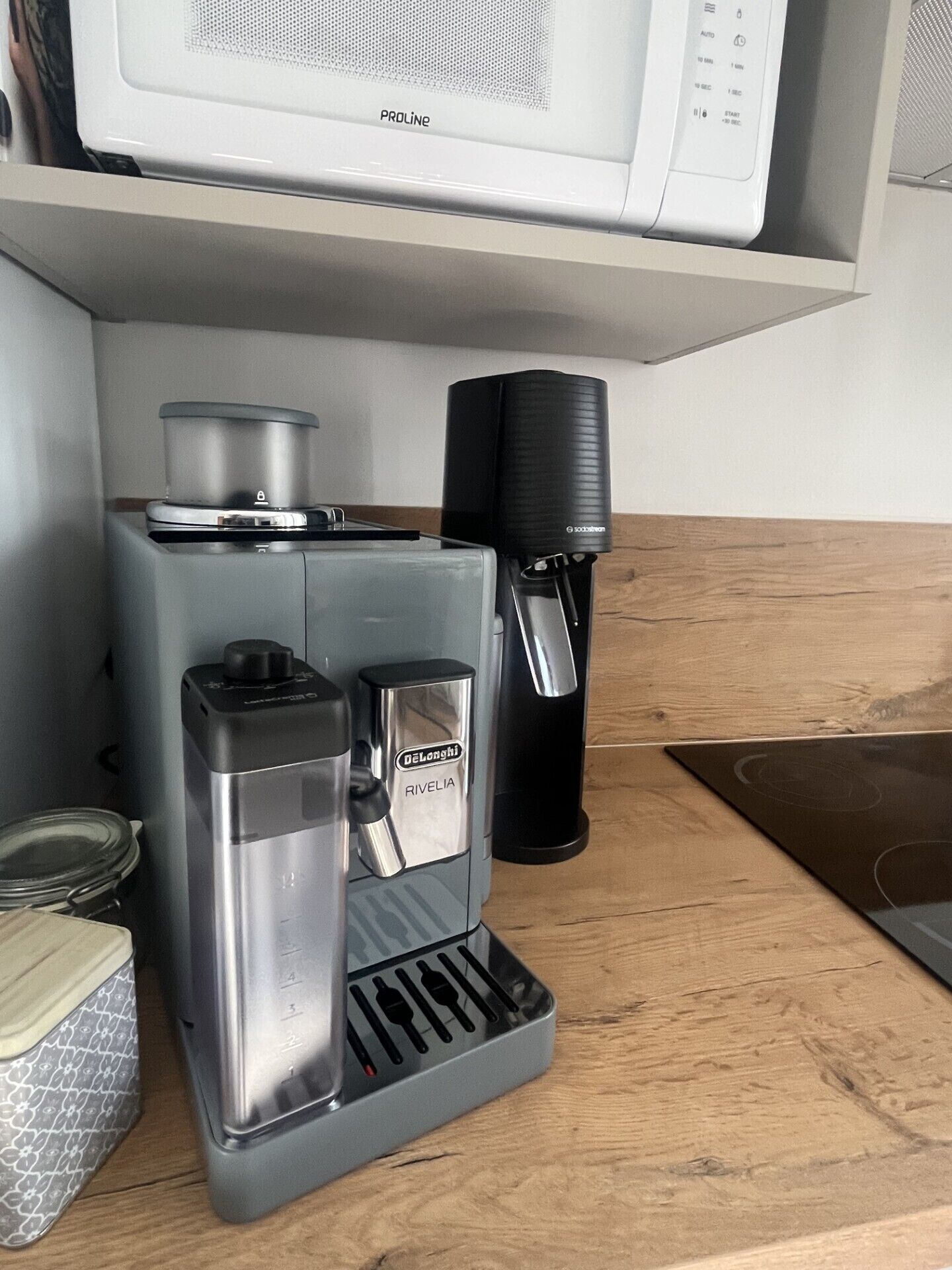Eine Kaffeemaschine der Marke Delonghi Rivelia, ein schwarzer Kaffeemühle und eine weiße Mikrowelle darüber.
