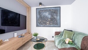 A modern living room with a large flat-screen TV, a round glass coffee table, a wooden console, a brown sofa with a green throw, and a large abstract painting on the wall. - Gîte confort de la Fontaine de Jonquier (Seguret)