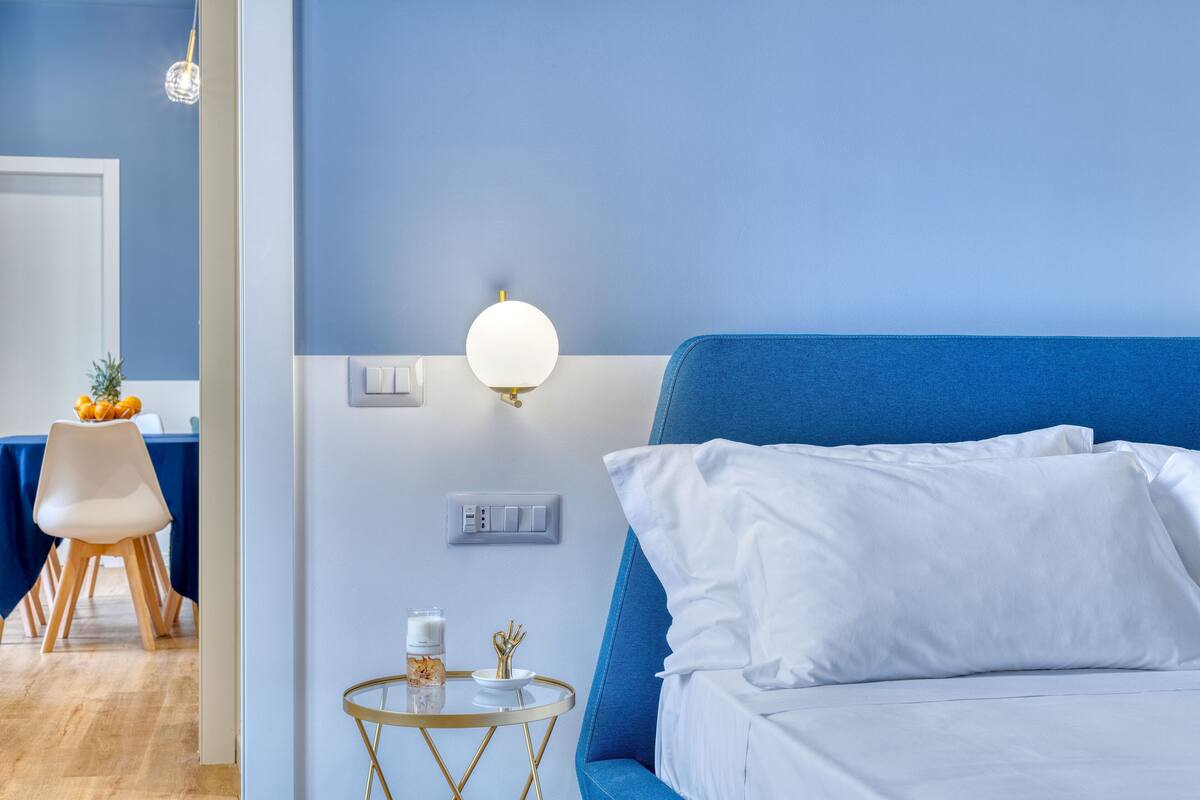 A bedroom with a blue headboard, white bedding, a bedside table with a lamp, and a wall socket.
