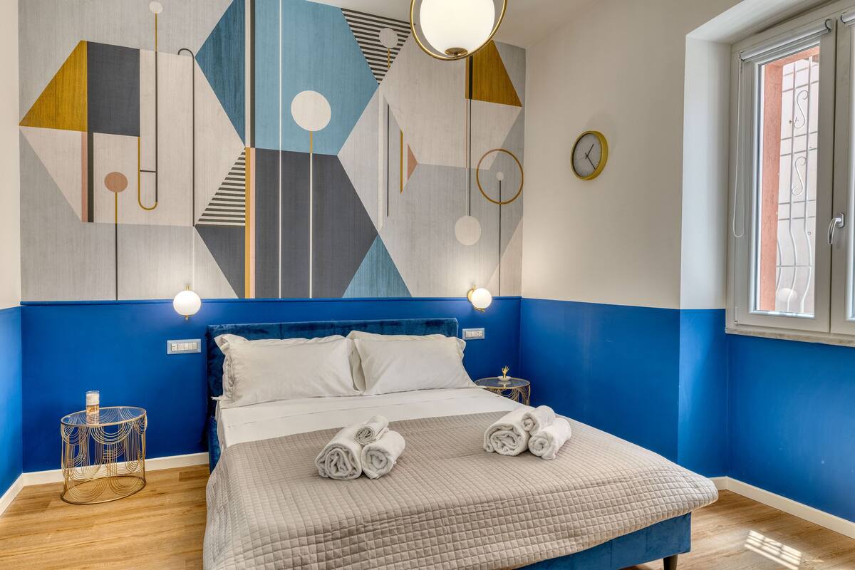 A modern hotel room with a blue and white color scheme, a large bed with a quilted bedspread, two bedside tables with lamps, and a geometric wall art piece.