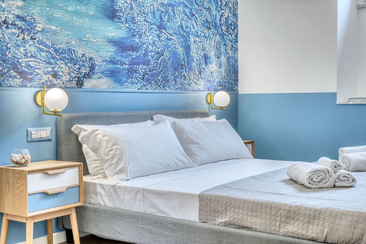 A neatly made bed with white linens, a bedside table with a drawer, and a wall-mounted light fixture.