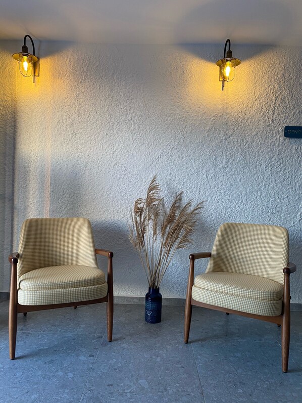 Two checkered armchairs with wooden legs and a vase with dried plants on a tiled floor against a textured wall with two wall-mounted lights. - Vahide Dalyan (Cesme)