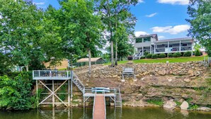 A wooden dock leading to a stone platform with a staircase, surrounded by lush greenery and a house in the background. - Allie’s on the Rocks Pet-Friendly Lakefront Bungalow (Cullman)