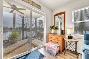 A room with a sliding glass door leading to a deck, a ceiling fan, a wooden dresser, a mirror, a lamp, and a plastic storage bin. - Allie’s on the Rocks Pet-Friendly Lakefront Bungalow (Cullman)