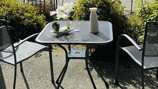 Un área de estar al aire libre con una mesa y sillas, un jarrón con flores, y una vista de un pueblo y agua al fondo.