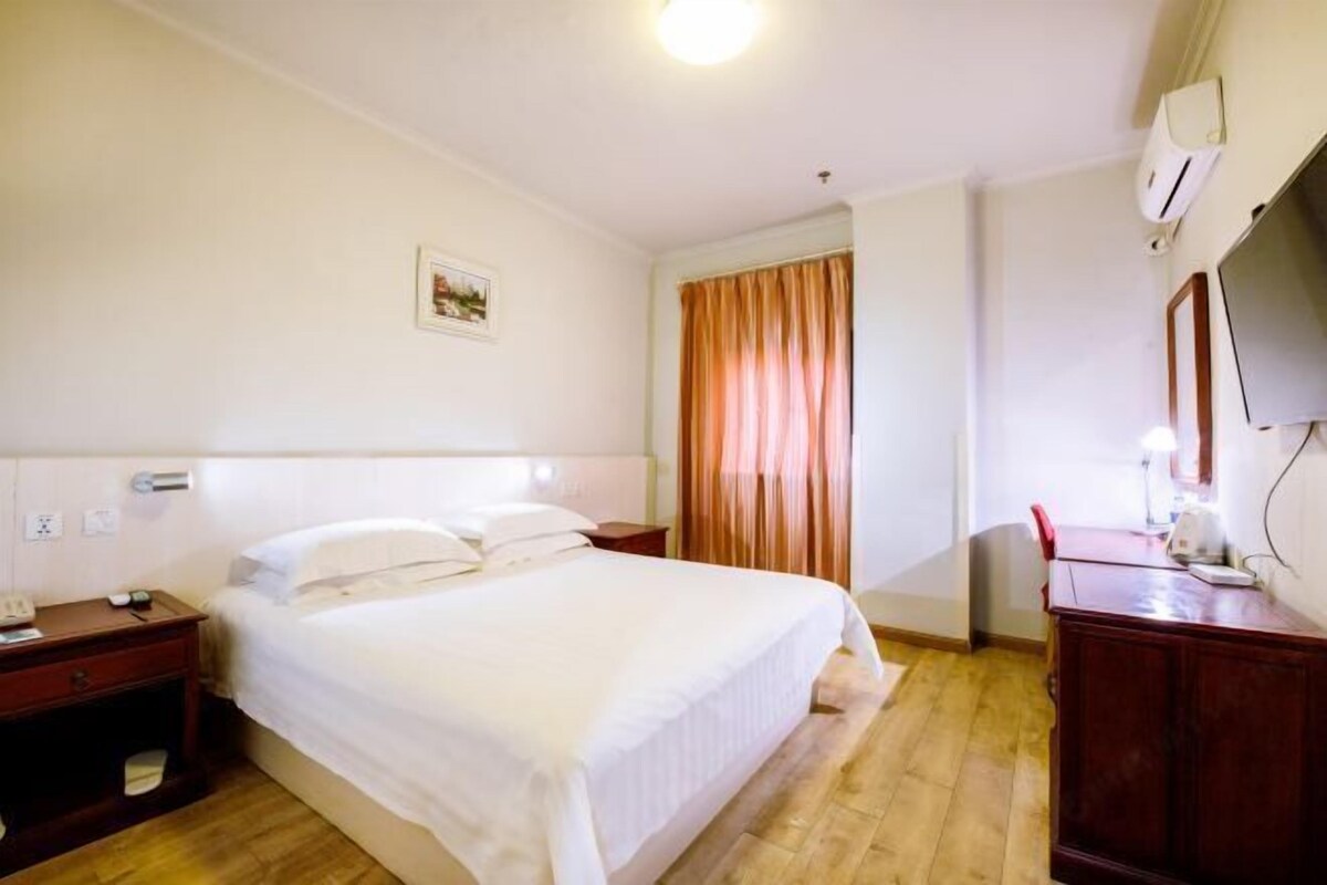 A hotel room with a single bed, a wooden nightstand, a desk with a chair, a television, and a window with curtains.