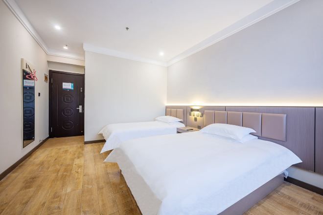 Quarto de hotel com duas camas, piso de madeira, uma porta e luminária fixada na parede.