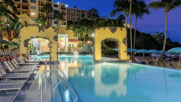 Ein Poolbereich eines Resorts mit Liegestühlen und Sonnenschirmen, umgeben von Palmen und einem Gebäude mit einer Steinaußenfassade.