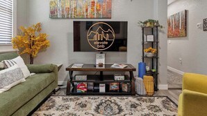 Un salon avec un canapé vert, un fauteuil jaune, un téléviseur, une bibliothèque et une plante décorative.