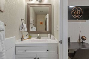 A bathroom with a white vanity, a large mirror, and a sink. There is a towel rack with white towels, a small potted plant, and a decorative item on the vanity. A television is mounted on the wall to the right. - Family friendly in Downtown Littleton (Littleton)