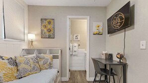 Une chambre à coucher avec un lit, un bureau et une chaise. Il y a un téléviseur accroché au mur et une porte qui donne sur une salle de bain.