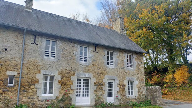 A stone building with multiple windows and a door, surrounded by autumn foliage. - Gîte Eloquent (Gratot)