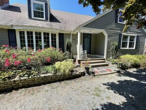 A two-story house with a stone foundation, a front porch with a white rocking chair, and a variety of flowering plants and shrubs.
