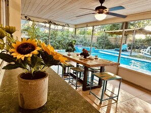 Un patio techado con vista a la piscina, una mesa con sillas y una planta de girasol grande.