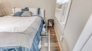 Un lit bien fait, avec une couette bleue et blanche, une petite table de chevet et un climatiseur fixé au mur.