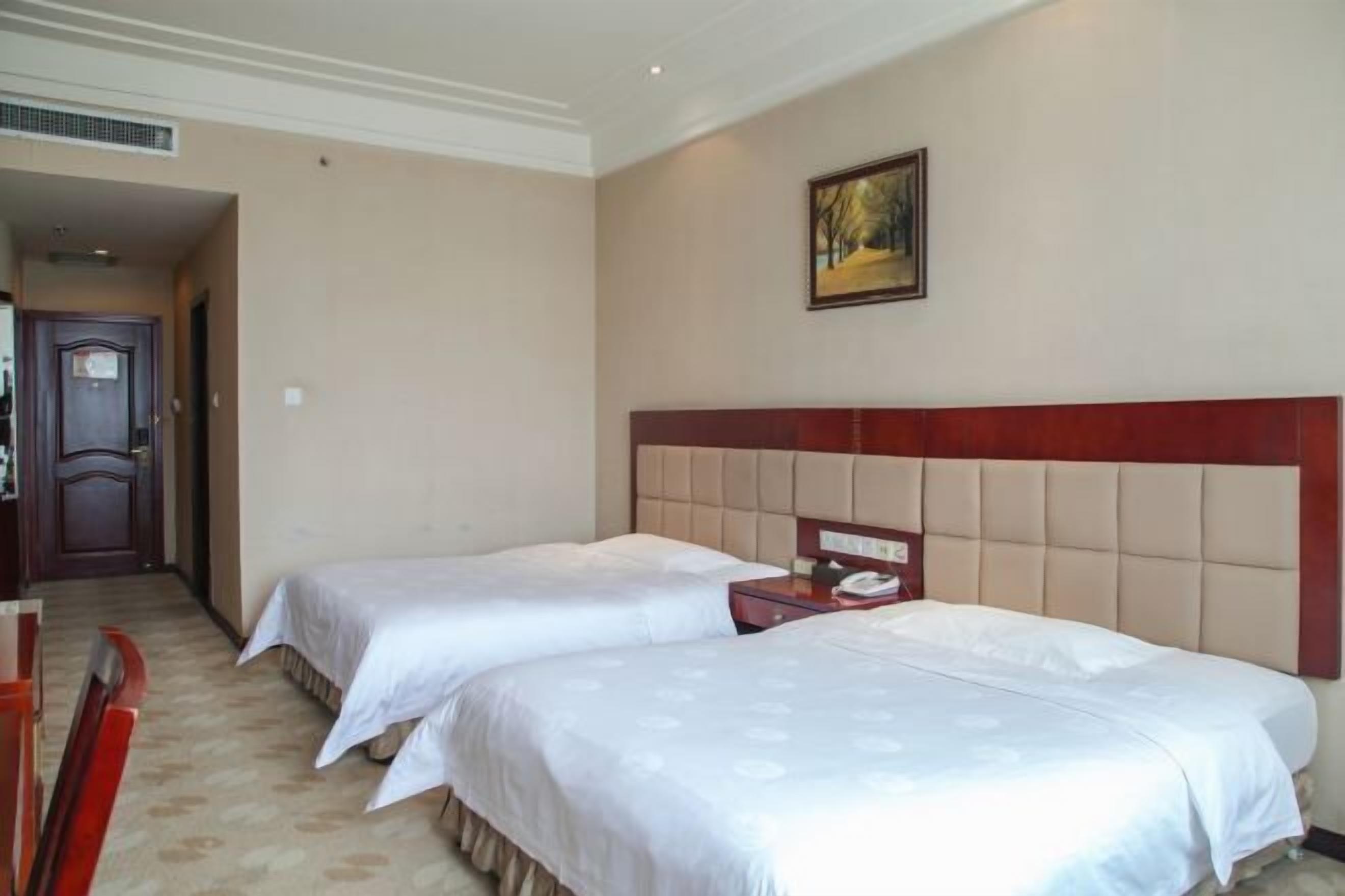 Habitación de hotel con dos camas, cabecero de madera, un cuadro en la pared y una puerta que da a otra habitación.