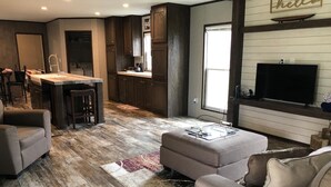 A modern kitchen with dark wood cabinets, a central island, and a bar area. There is a large window providing natural light and a flat-screen TV mounted on the wall. - Relax, Enjoy, Explore, On Cane! (Natchez)