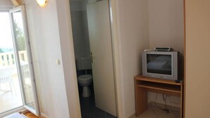Una habitación con un inodoro, un televisor sobre un pedestal de madera, y una puerta que da a un balcón.