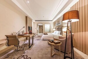 Habitación de hotel con escritorio, silla, cama, mesita de noche, lámpara y vista a otra habitación.