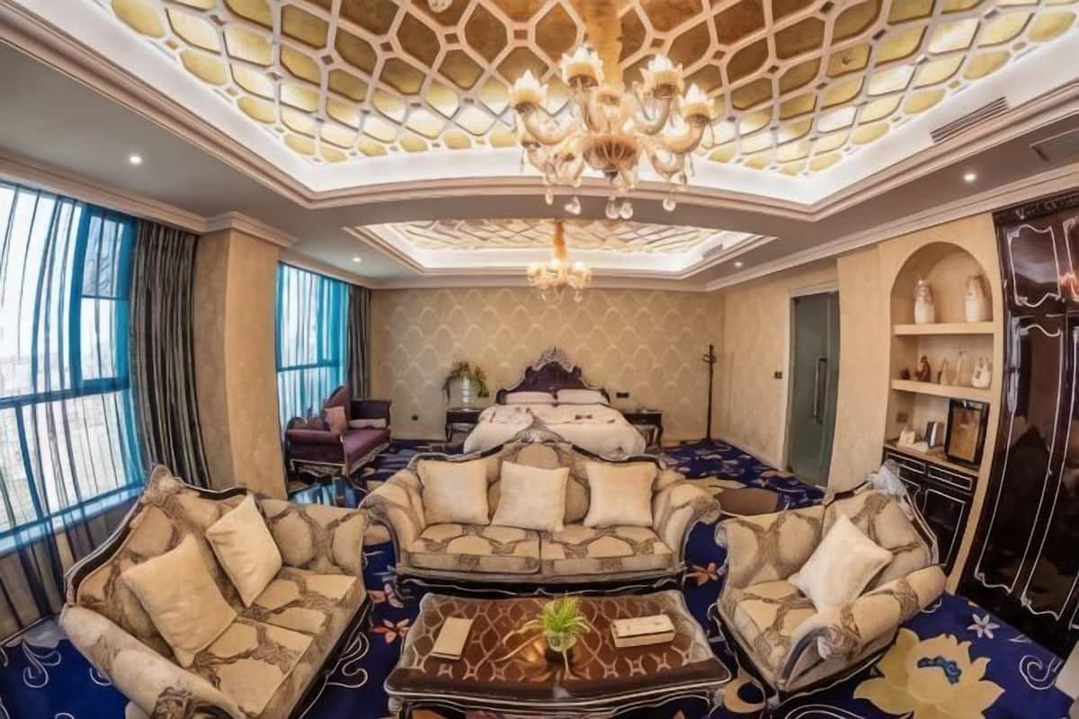 Una habitación lujosa con techo alto, una araña, una cama grande y una zona de estar con sofás y una mesa de centro.