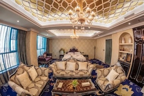 Une chambre luxueuse avec un plafond haut, un lustre, un grand lit et un coin salon comprenant des canapés et une table basse.