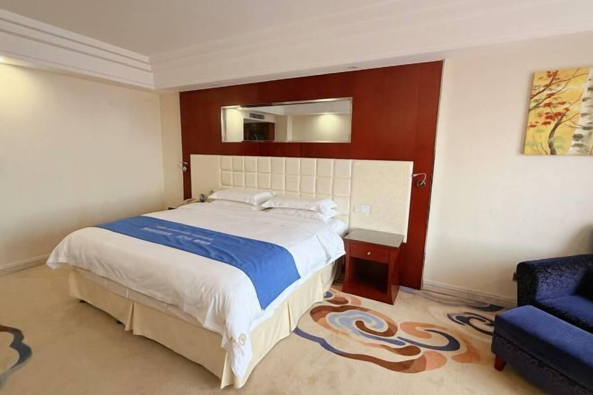 Habitación de hotel con una cama grande, un cabecero con espejo, una mesita de noche pequeña, una silla azul y una alfombra decorativa.