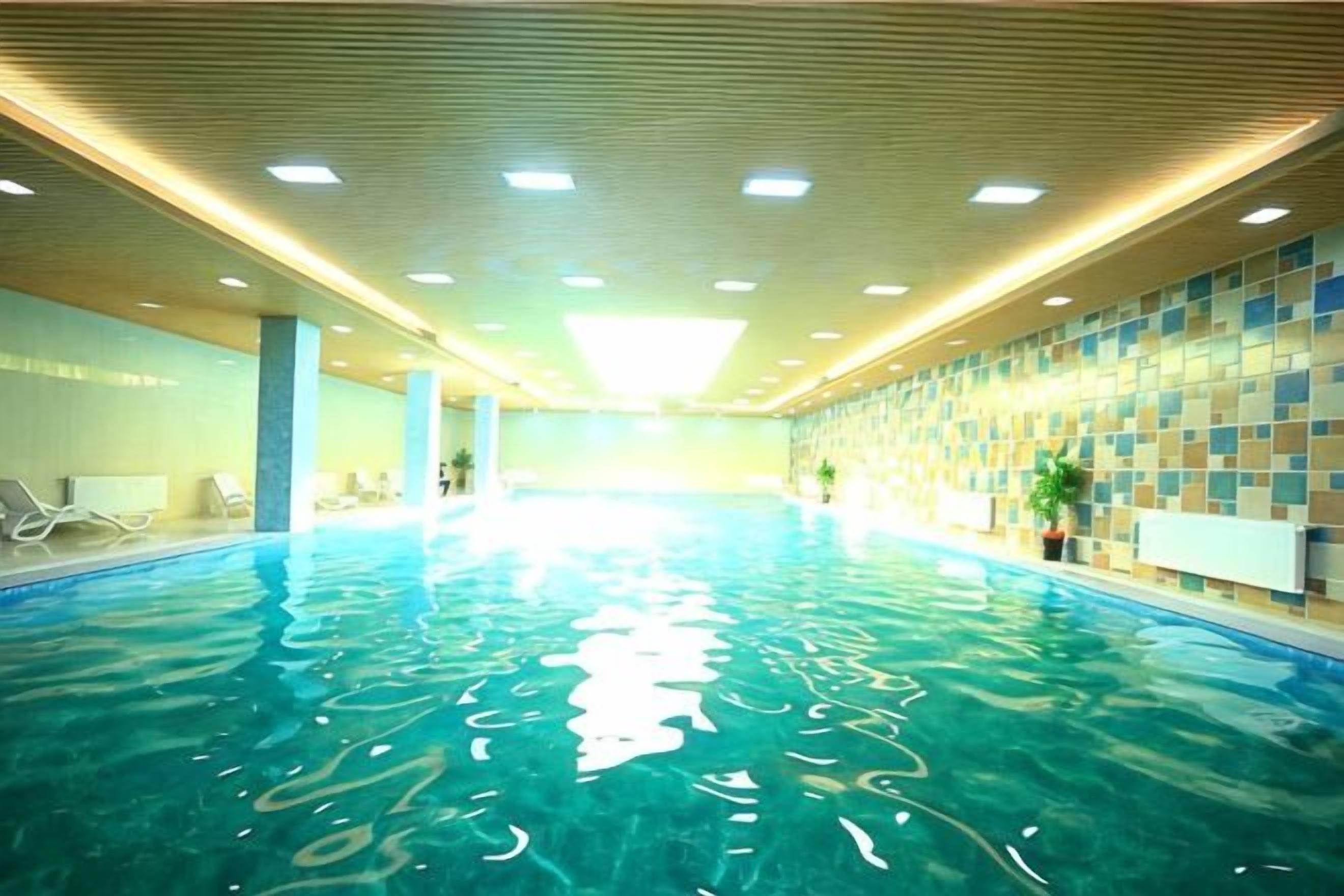 An indoor swimming pool with clear water, surrounded by walls with a patterned design, and a ceiling with recessed lighting.
