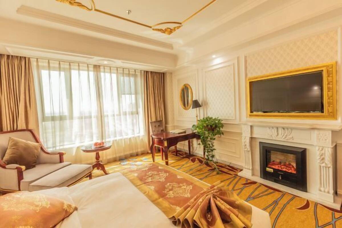 Habitación de hotel con cama, sofá, escritorio, televisor, chimenea y un ventanal grande con cortinas.