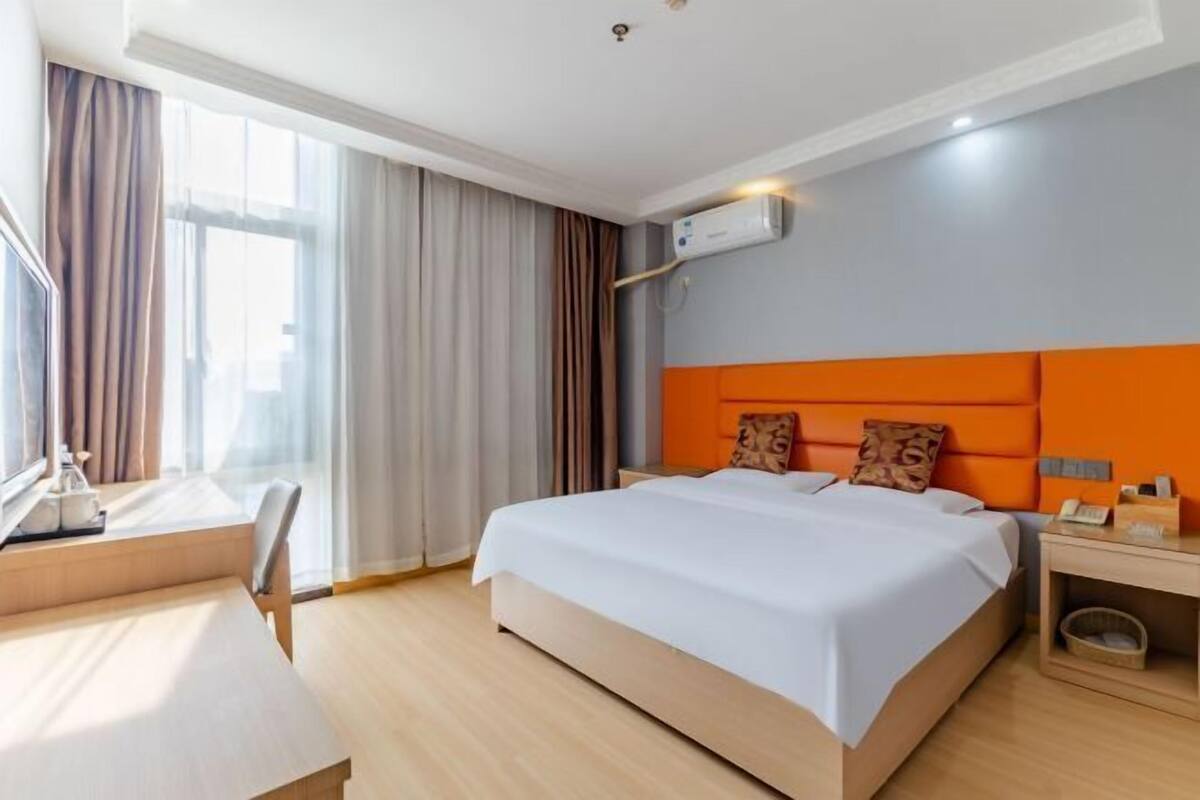 A hotel room with a bed, a desk, a chair, a bedside table, and an air conditioning unit.