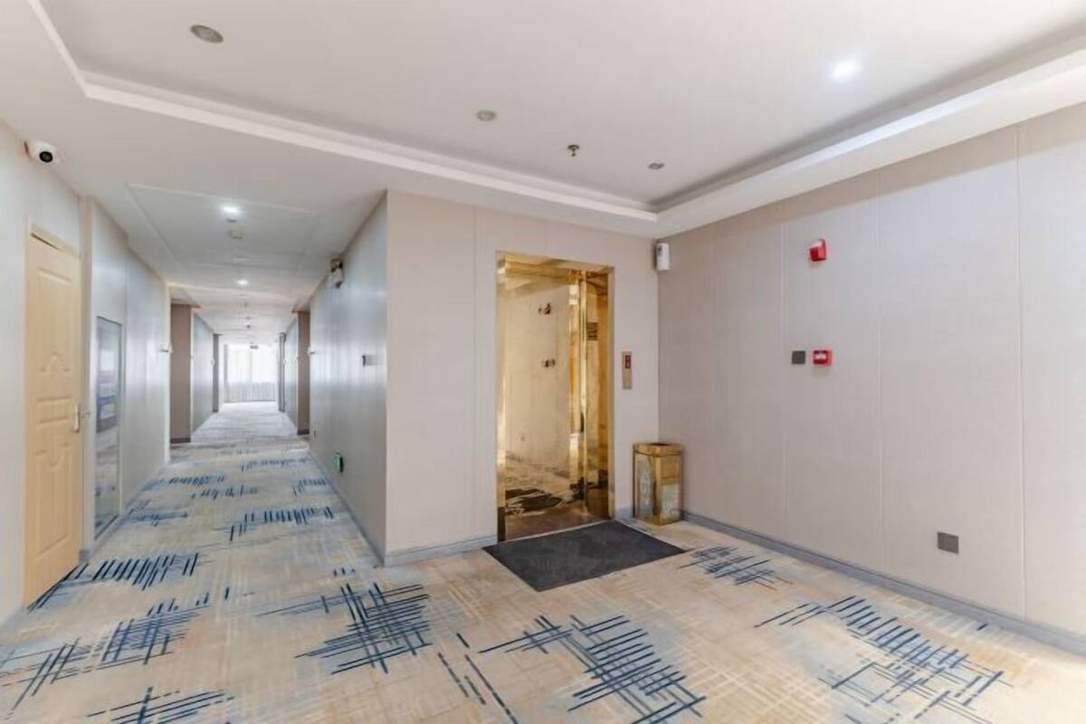 A hotel corridor with a patterned floor, a door on the left, and a wall-mounted fire alarm on the right.
