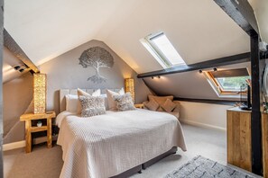 A bedroom with a sloped ceiling, a bed with multiple pillows, a nightstand with a lamp, and a wooden dresser.