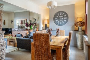 A dining area with a wooden table, woven chairs, a large wall clock, and a fireplace.