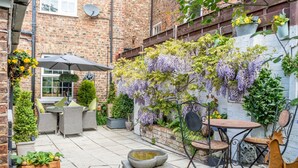 Ein Ziegelgebäude mit einer Terrassenfläche, die Gartenmöbel, eine hängende Blumenattrappe und einen kleinen Garten zeigt.